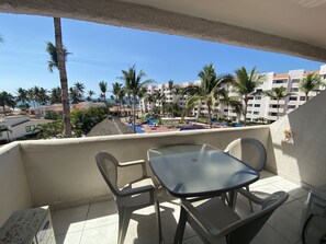 A balcony with a table and chairs, overlooking a pool area with palm trees and buildings in the background.
