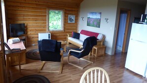Puinen mökki, jonka sisätiloissa on sohva, kaksi nojatuolia, televisio, jääkaappi ja taulu seinällä.