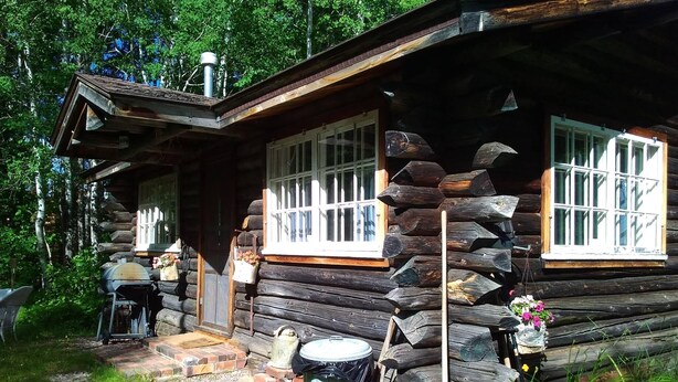 Une cabane rustique en rondins de bois, dotée d’une cheminée, d’un porche et d’un barbecue à l’extérieur.