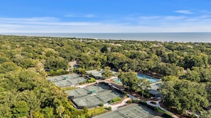 An aerial view of a residential area with multiple tennis courts, surrounded by dense greenery and a view of the sea.