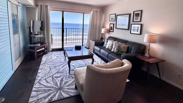 Una sala de estar con un sofá, un sillón y una mesa de centro. Hay un televisor sobre un mueble bajo, una lámpara y un cuadro enmarcado en la pared. Un amplio ventanal ofrece vistas a la playa.