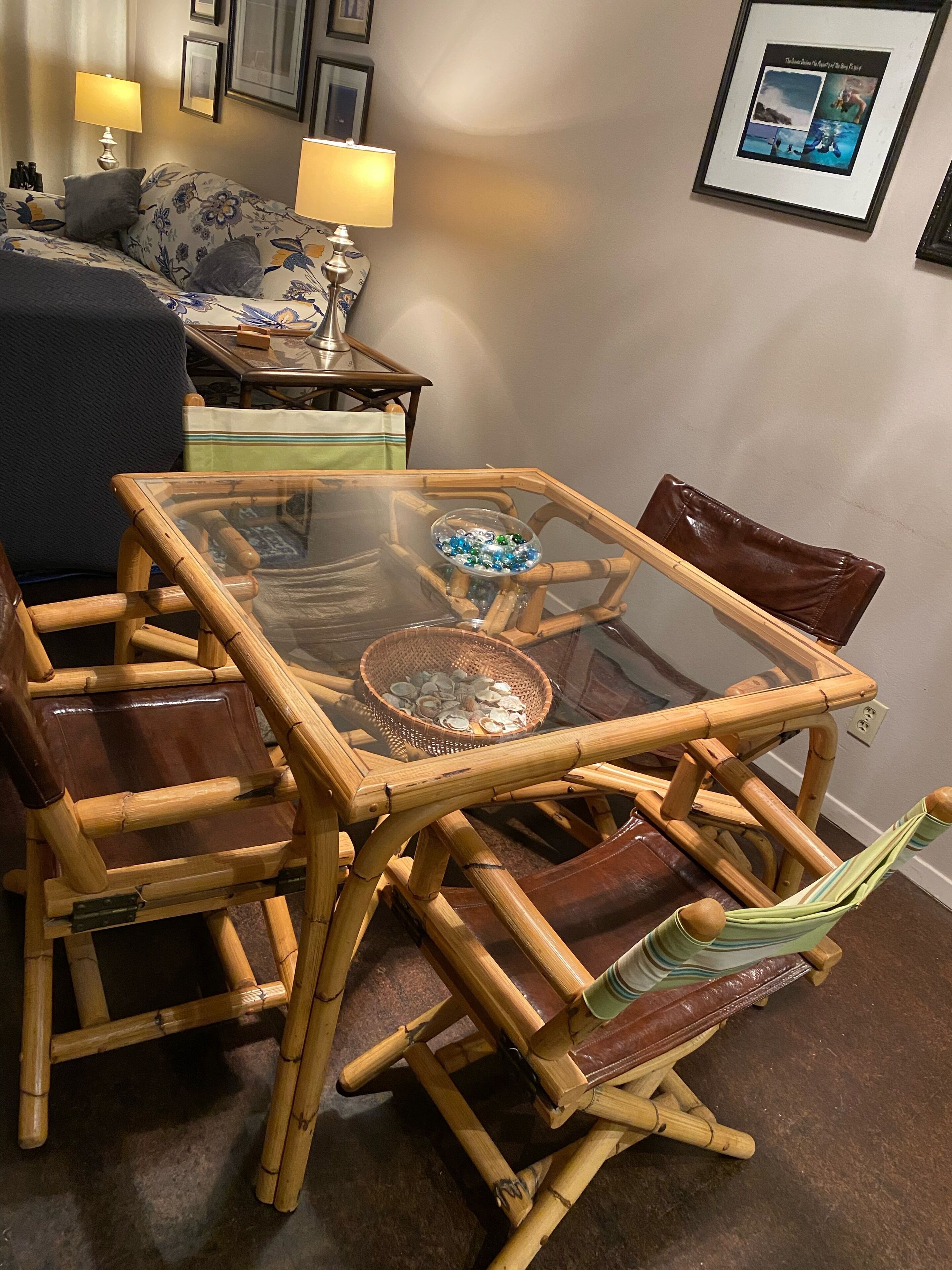 Una mesa de comedor de bambú con superficie de vidrio, rodeada de sillas de cuero, y una cesta de conchas marinas sobre la mesa.