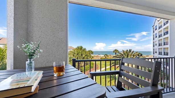 Ein Balkon mit Blick auf das Meer, ein Tisch mit einem Buch und einem Getränk sowie ein Stuhl.