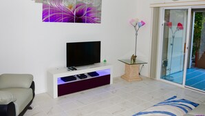 A living room with a flat-screen TV on a white entertainment unit, a beige sofa, a glass coffee table, and a large sliding glass door leading to an outdoor area. - Mid-Term Stays in Gorgeous Condo
 Kona, Big Island (Kailua-Kona)