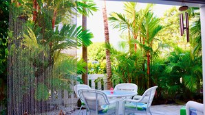 An outdoor patio with wicker furniture and a table, surrounded by lush greenery and palm trees. - Mid-Term Stays in Gorgeous Condo
 Kona, Big Island (Kailua-Kona)