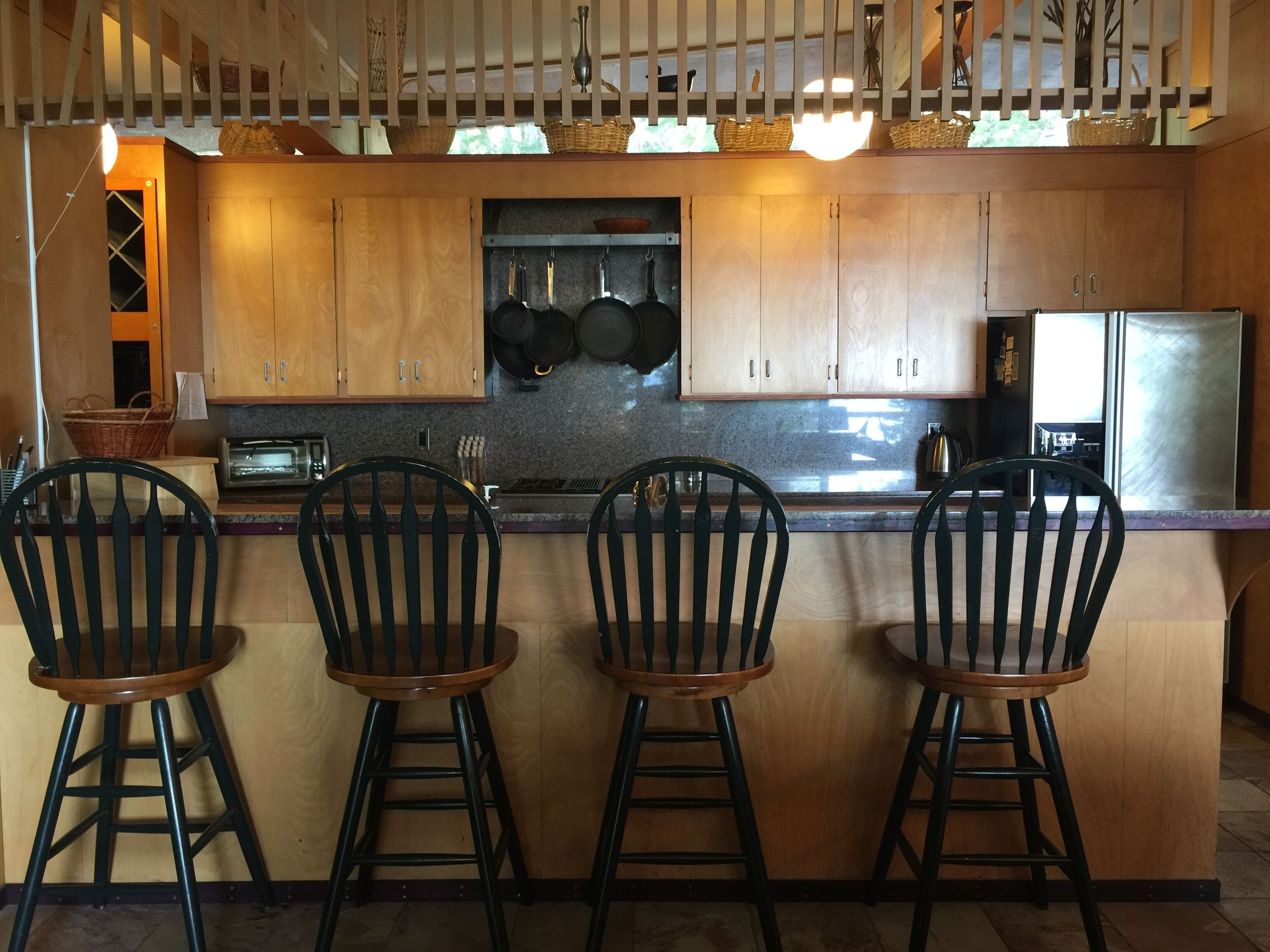 A kitchen with wooden cabinets, a granite countertop, and a stainless steel refrigerator. There are four bar stools with wooden seats and metal legs.