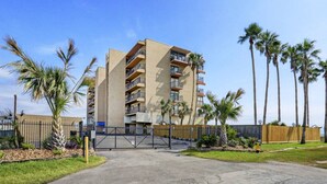A multi-story building with balconies, surrounded by palm trees and a gated entrance.
