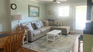 A living room with a sofa, coffee table, and chairs. There is a ceiling fan, a clock, and a window with blinds.