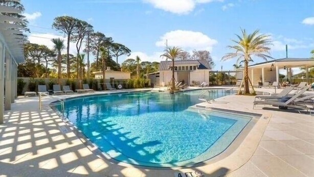 A swimming pool with sunlit areas and lounge chairs, surrounded by palm trees and a building with a sloped roof.