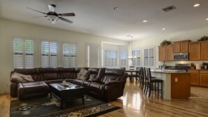 A spacious living room with a leather sectional sofa, a dining area with a kitchen in the background, and large windows with blinds. - 2400 sq. ft., 4 bedroom, 2 bath, sleeps 8, hot tub (Goodyear)
