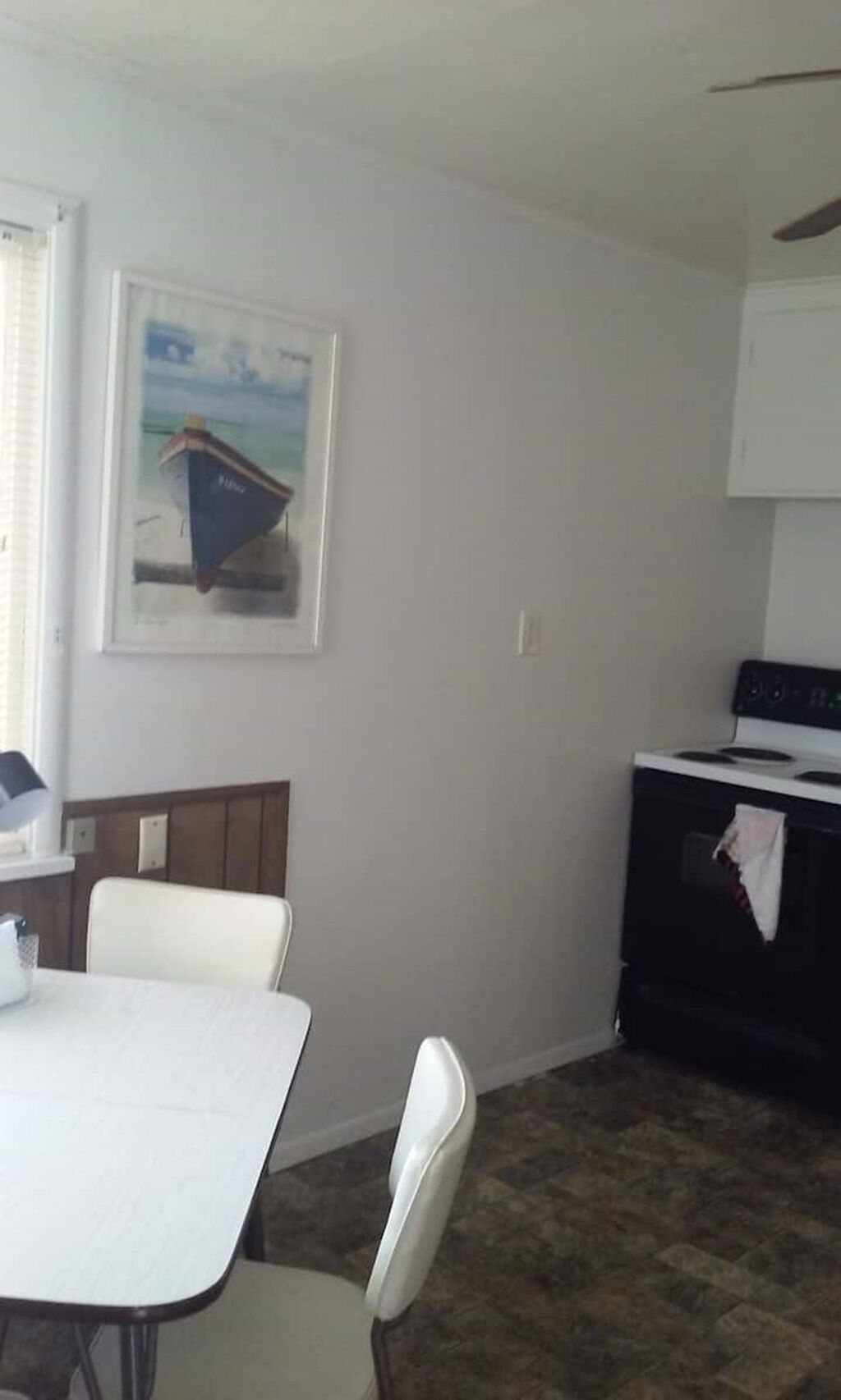 Una habitación pequeña y poco amoblada con una mesa blanca, una silla blanca, un cuadro con una imagen de un barco, una estufa y un piso de baldosas.