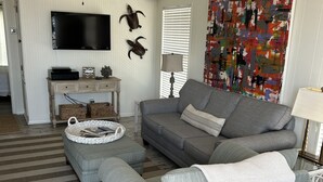A living room with a grey sofa, a grey ottoman, a ceiling fan, a flat-screen TV, and a colorful abstract painting. - East End -Beach Oasis- St. George Island. Beach Condo / Full Sized Swimming Pool (St.George Island)