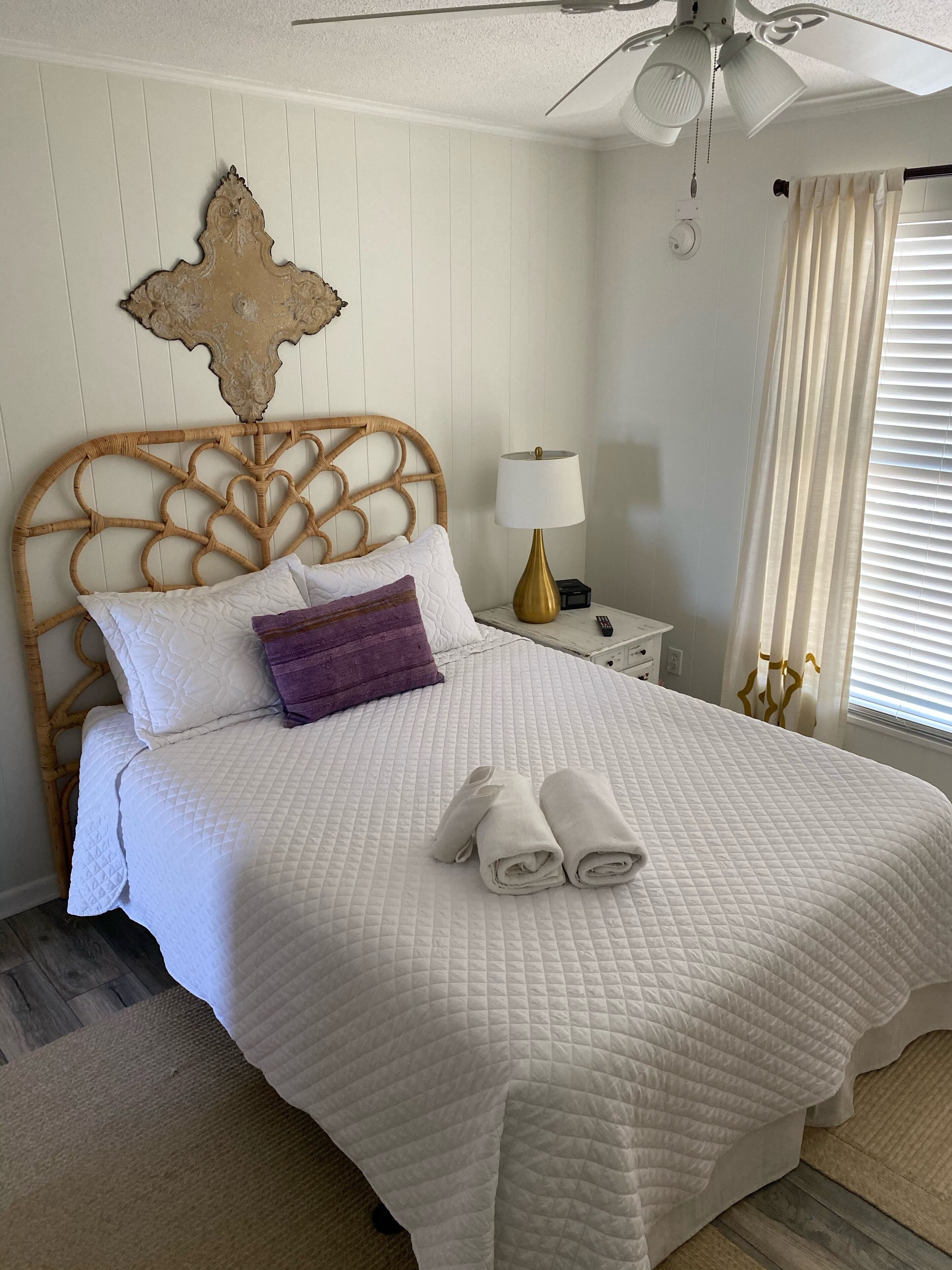 A neatly made bed with a white quilt, a purple pillow, and two rolled towels.