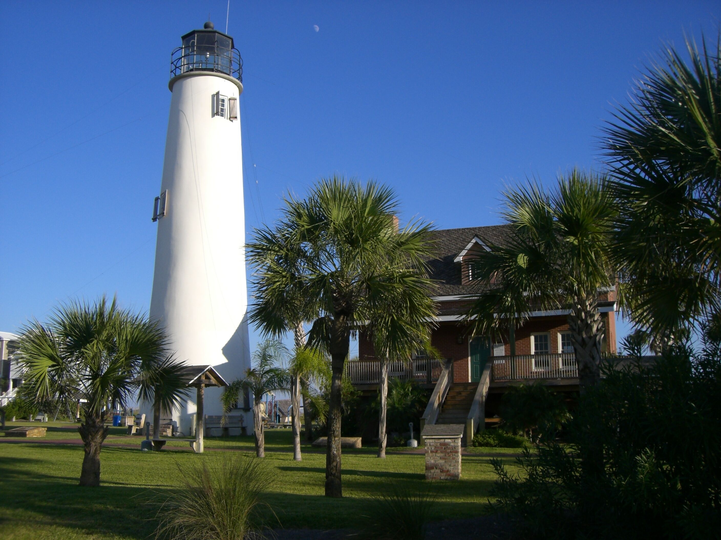 A lighthouse with a white cylindrical tower, surrounded by palm trees and a brick building with a balcony.