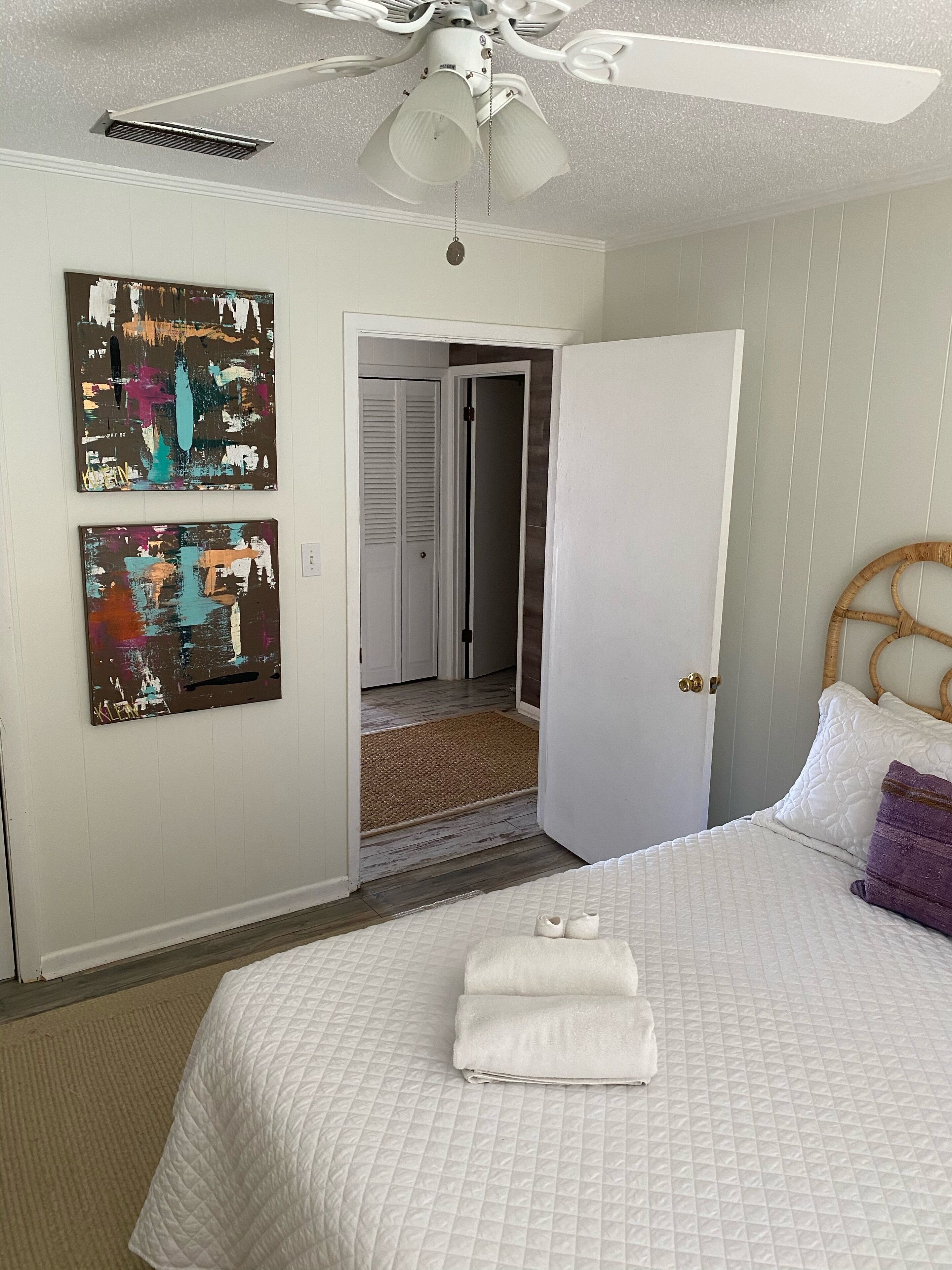 A bedroom with a bed, a ceiling fan, two abstract paintings, and a door leading to a hallway.