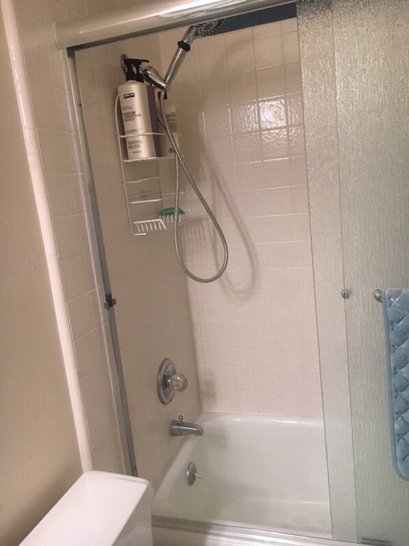 A shower area with a glass door, a handheld showerhead, a wall-mounted soap dispenser, and a white bathtub.