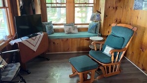A cozy cabin interior with a wooden floor, a television, a rocking chair, a bench, and a window seat with a view of trees. - Peaceful Lakefront Retreat with Views (Newfield)