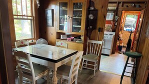 A wooden dining table with chairs, a kitchen area with cabinets and appliances, and a view of the outdoors through a window. - Peaceful Lakefront Retreat with Views (Newfield)