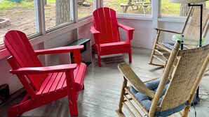A cozy room with large windows, red Adirondack chairs, a wicker rocking chair, and a table. - Peaceful Lakefront Retreat with Views (Newfield)