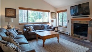 Un salón con sofá, sillón, mesa de centro de madera, chimenea, televisor y ventanas amplias.
