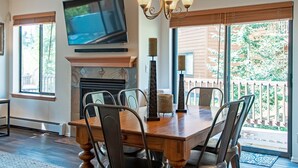 Un comedor con mesa y sillas de madera, una chimenea, un televisor montado en la pared y un balcón con vistas a árboles.