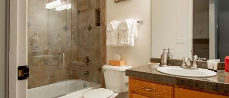 A bathroom with a glass-enclosed shower, a white toilet, a sink with a granite countertop, wooden cabinets, and a mirror.