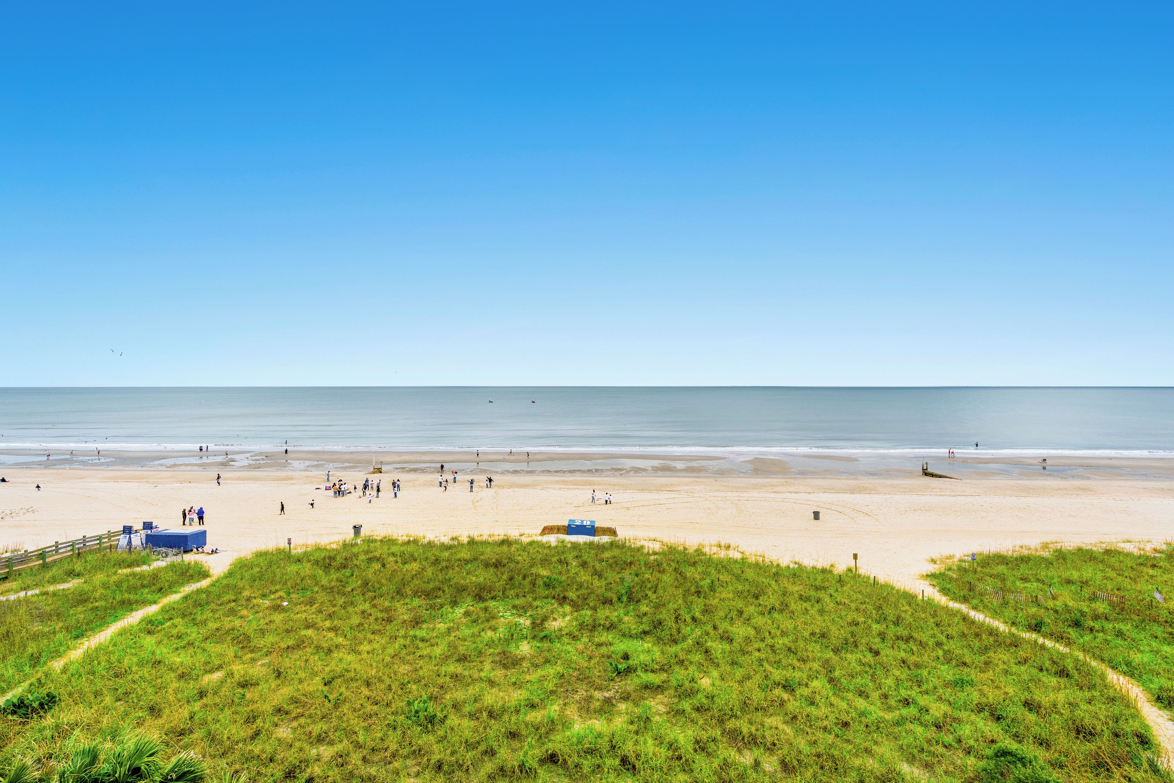 A beach with a clear sky, grassy dunes, and a sandy shore with people and a lifeguard tower.