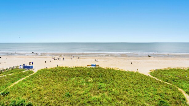 A beach with a clear sky, grassy dunes, and a sandy shore with people and a lifeguard tower. - Oceanfront Condo with indoor pool + Free Attraction Tickets! (North Myrtle Beach)