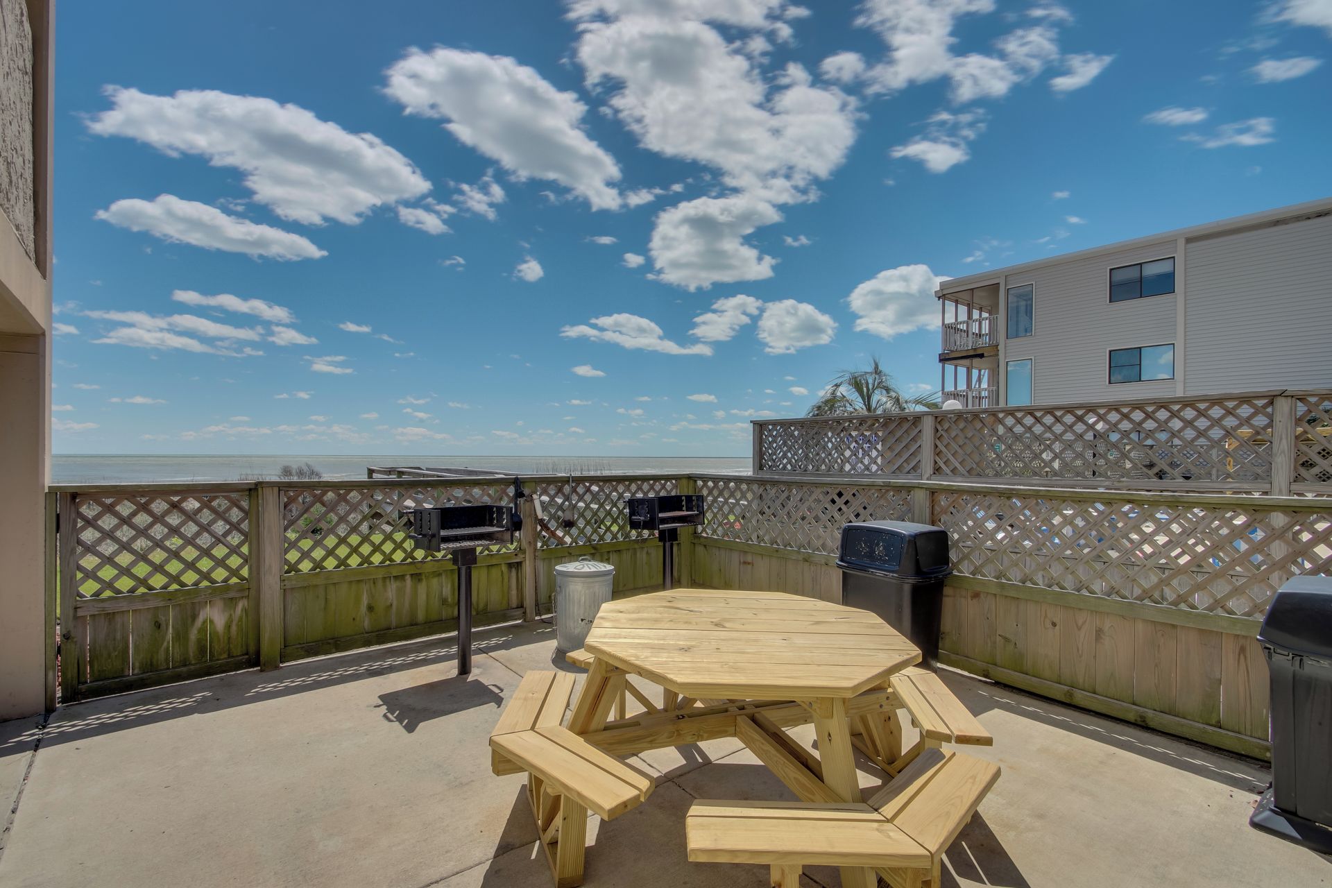 A wooden deck with picnic tables and barbecue grills, overlooking a body of water and a building with balconies.