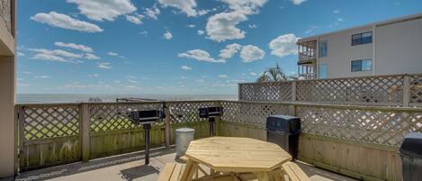 A wooden deck with picnic tables and barbecue grills, overlooking a body of water and a building with balconies.