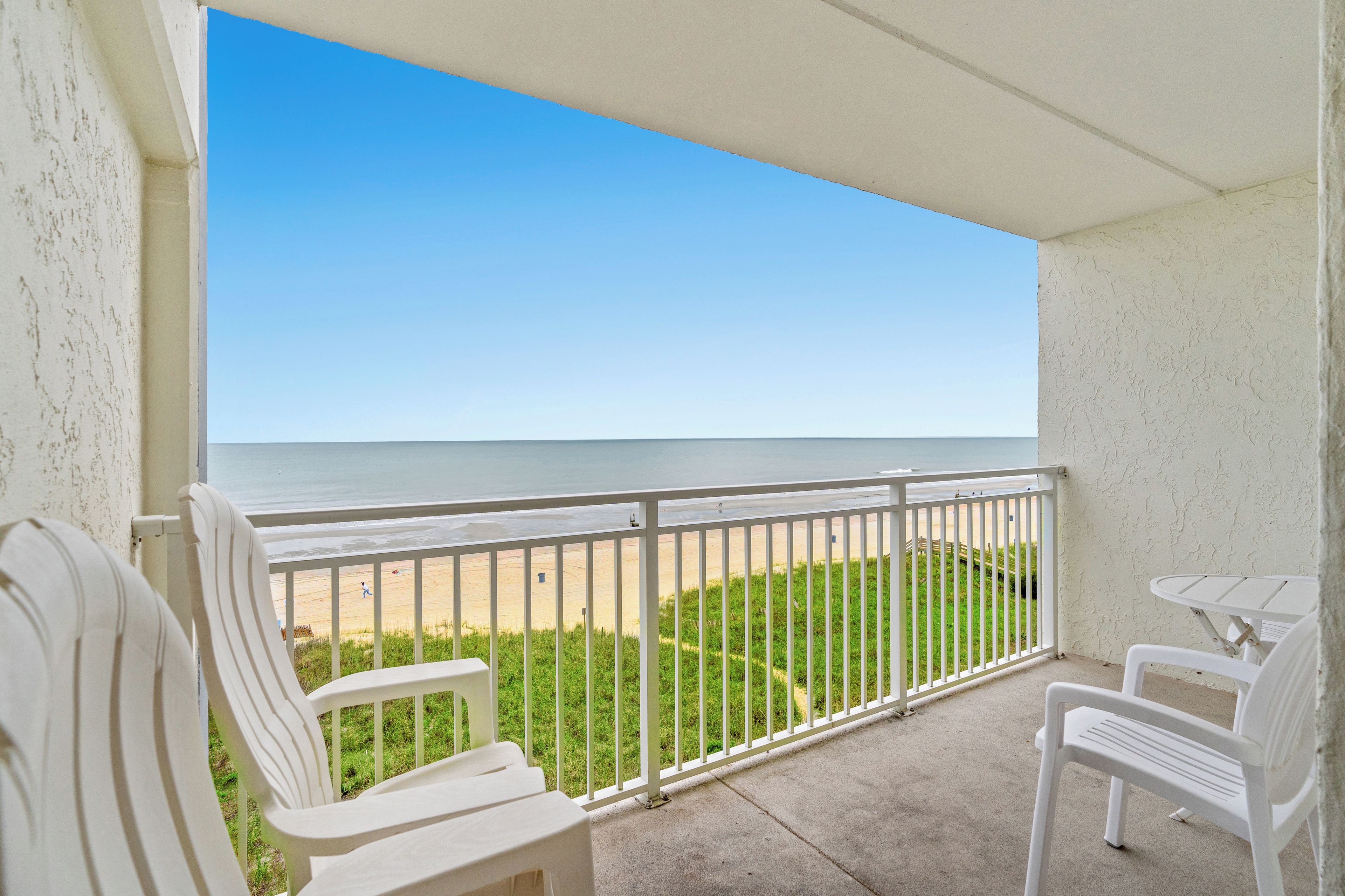 A balcony with white chairs and a table, overlooking a beach with a clear sky.