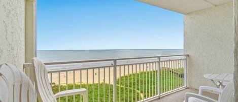 A balcony with white chairs and a table, overlooking a beach with a clear sky.