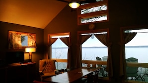 Een kamer met een groot raam met uitzicht op een wateroppervlak, een plafondventilator, een lamp, een stoel en een tafel.
