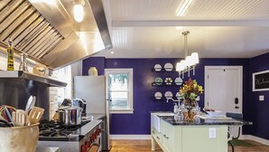 Une cuisine équipée d’une cuisinière, d’un réfrigérateur et d’un îlot central aux couleurs claires. On y trouve des assiettes décoratives au mur et une fenêtre offrant une belle vue.