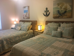 Une chambre avec deux lits, une table de chevet avec une lampe et des œuvres d’art murales représentant une scène de plage.