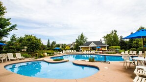 Uma grande piscina ao ar livre com várias áreas, rodeada por espreguiçadeiras brancas e guarda-sóis azuis.