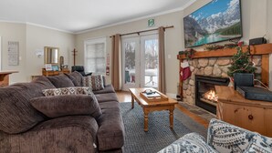 Uma sala de estar acolhedora com lareira, uma janela ampla com vista para montanhas cobertas de neve e um sofá confortável.