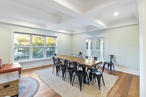 Uma sala de jantar com mesa de madeira e cadeiras pretas, grandes janelas e vista para árvores.
