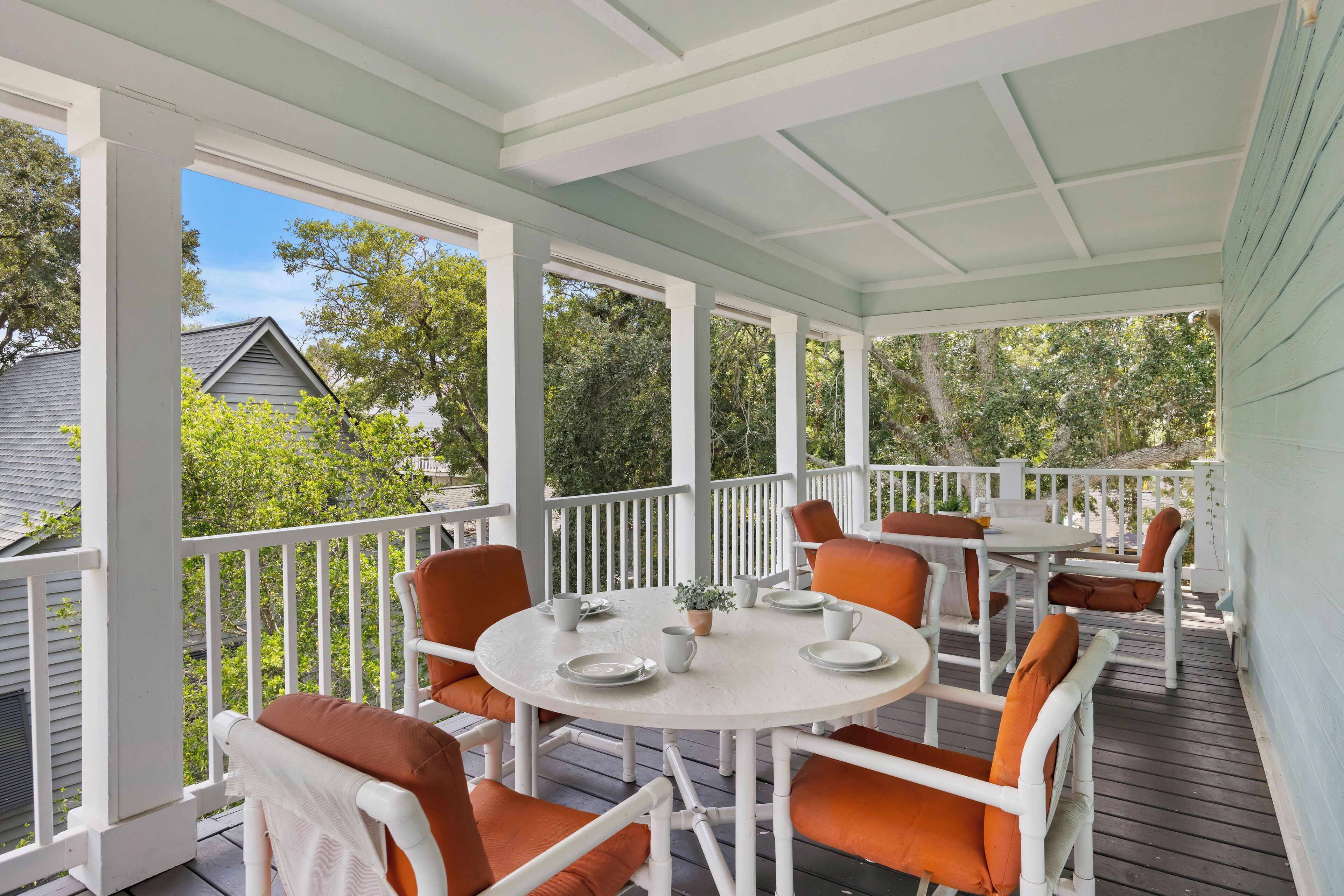 A covered outdoor seating area with white and orange chairs, a round table, and a view of trees and a house in the background.