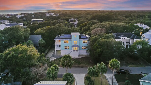 Een groot huis met een blauw-wit uiterlijk, omringd door bomen en een uitzicht op de oceaan bij zonsondergang.