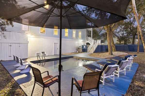 Un área de piscina con sillas de descanso y una sombrilla, rodeada por una casa con escaleras y una valla.