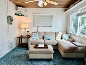 A cozy living room with a sectional sofa, a side table, a lamp, and a window with blinds. - Pass-a-Grille Gulf Front Cottage, Ocean View (St Pete Beach)