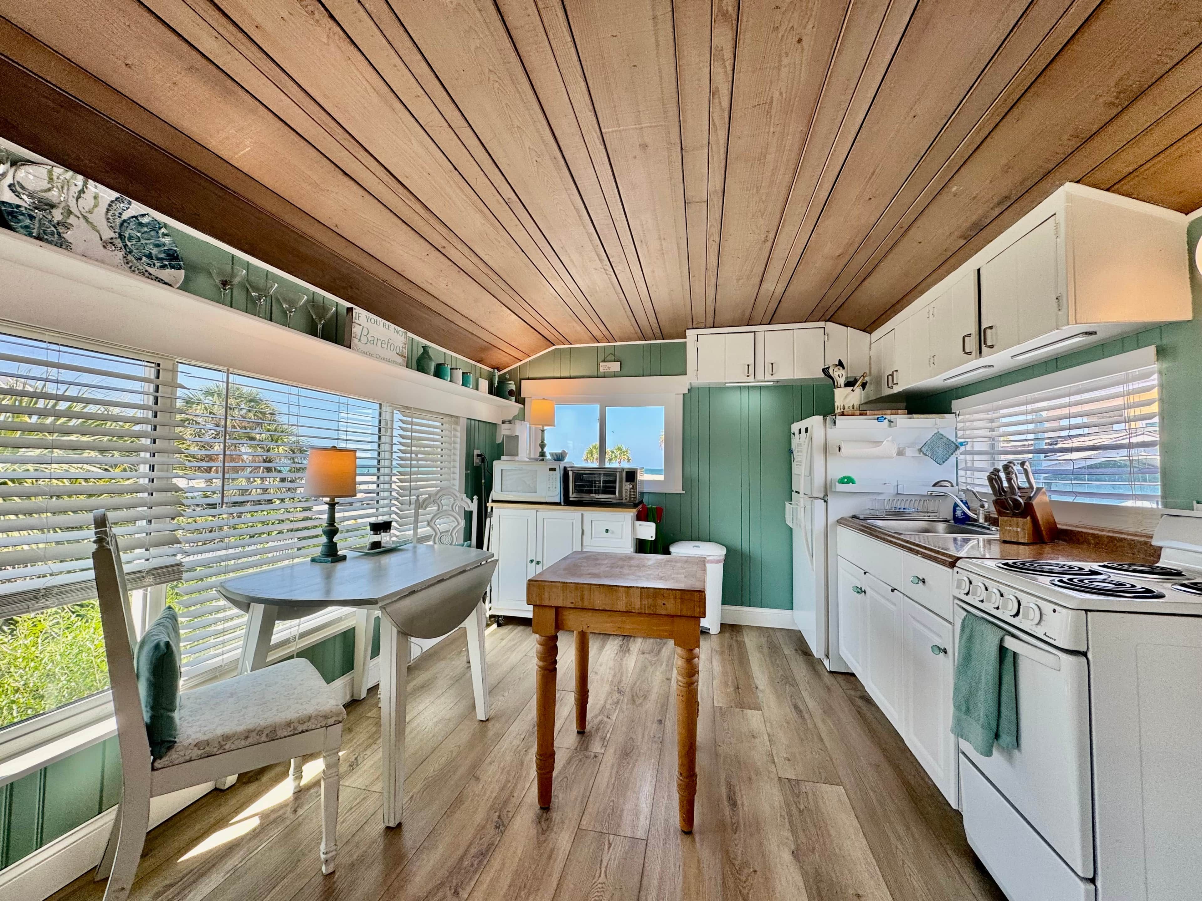 A kitchen with wooden ceiling, white cabinets, a dining table with chairs, and a window with blinds.
