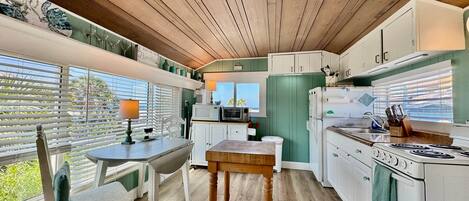 A kitchen with wooden ceiling, white cabinets, a dining table with chairs, and a window with blinds.