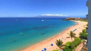 A beach with clear waters, a sailboat, and a resort area with buildings and palm trees.