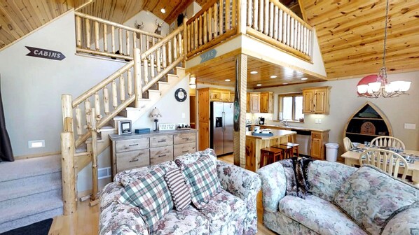 A cabin interior with a wooden staircase, a kitchen, a dining area, and a living room with floral-patterned sofas.