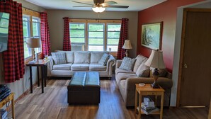 Ein Wohnzimmer mit einem beigen Sofa, einem Couchtisch aus Holz und einem Deckenventilator. Zwei Fenster mit roten und schwarzen Karottenvorhängen und ein Gemälde an der Wand.