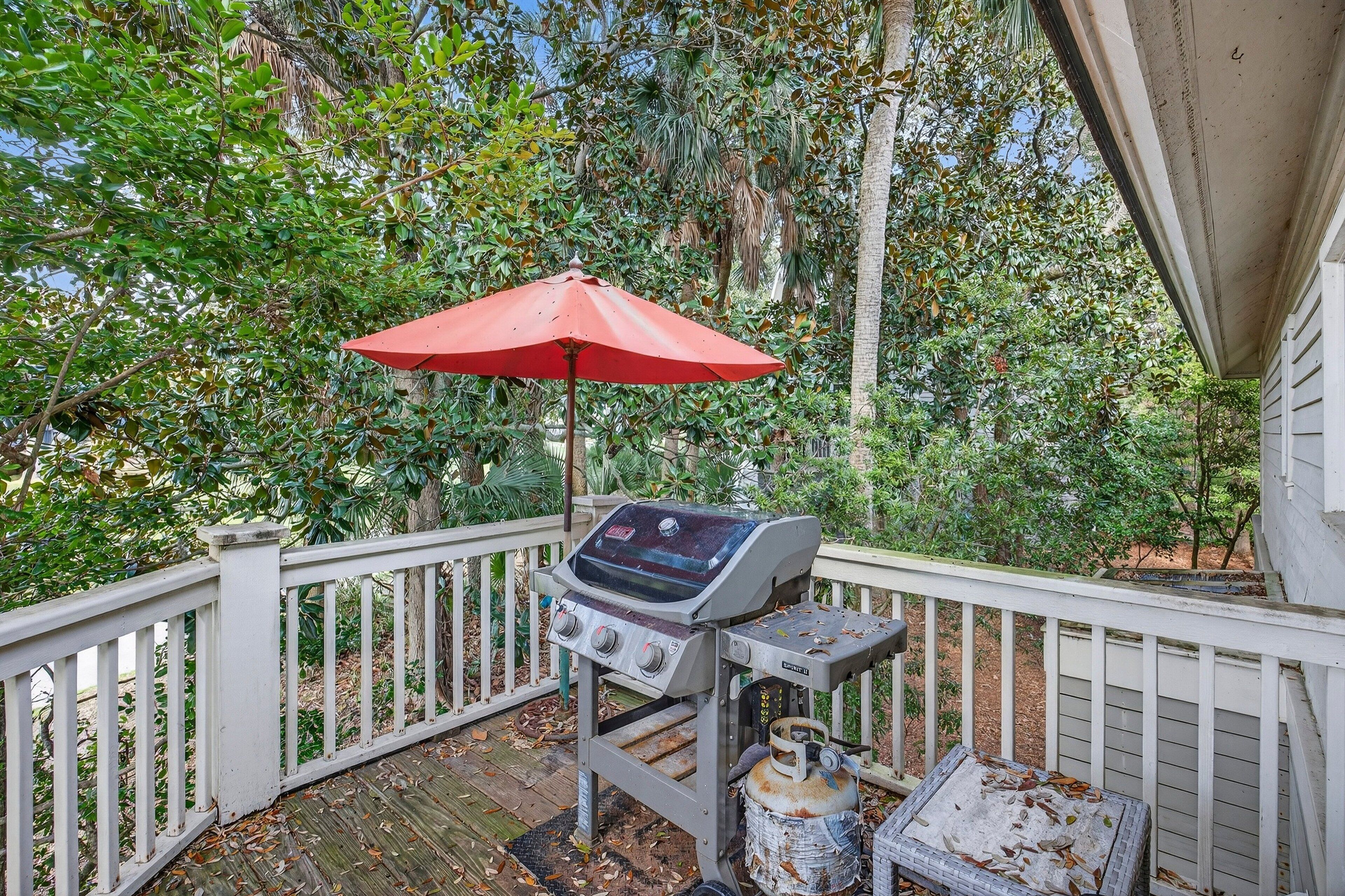 Un barbecue installé sur une terrasse en bois dotée d’une rambarde blanche, le tout entouré d’une végétation luxuriante.