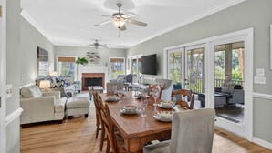 Uma ampla área de jantar com uma mesa de madeira posta para refeição, cercada por assentos confortáveis. Um ventilador de teto e uma lareira são visíveis ao fundo.