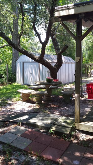 A garden with a wooden pergola, a table with a watermelon, and a shed in the background. - Turtle Time - 4 Bd House Near Holden Beach - escape the crowds! (Supply)
